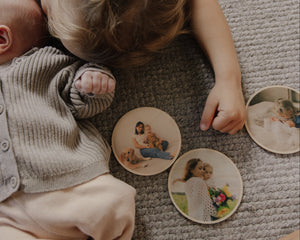 A baby and toddler lying on a textured gray carpet, interacting with round wooden coasters printed with family photographs.